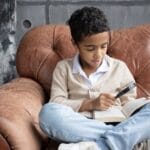 Full body of curious Arabian schoolboy sitting on comfortable armchair with crossed legs and reading book with magnifier at home