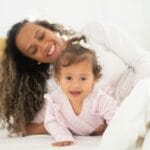 A joyful mother and baby daughter enjoy a loving moment on a comfy bed with natural light.