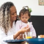A loving mother feeds her young daughter breakfast at home, emphasizing care and togetherness.