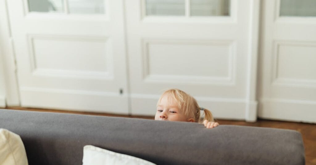 A blonde child playfully peeks over a gray sofa in a cozy living room setting.