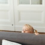 A blonde child playfully peeks over a gray sofa in a cozy living room setting.