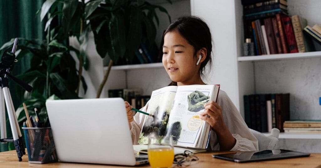 Asian child studying at home with a book and laptop, engaged in video calling.