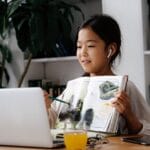 Asian child studying at home with a book and laptop, engaged in video calling.