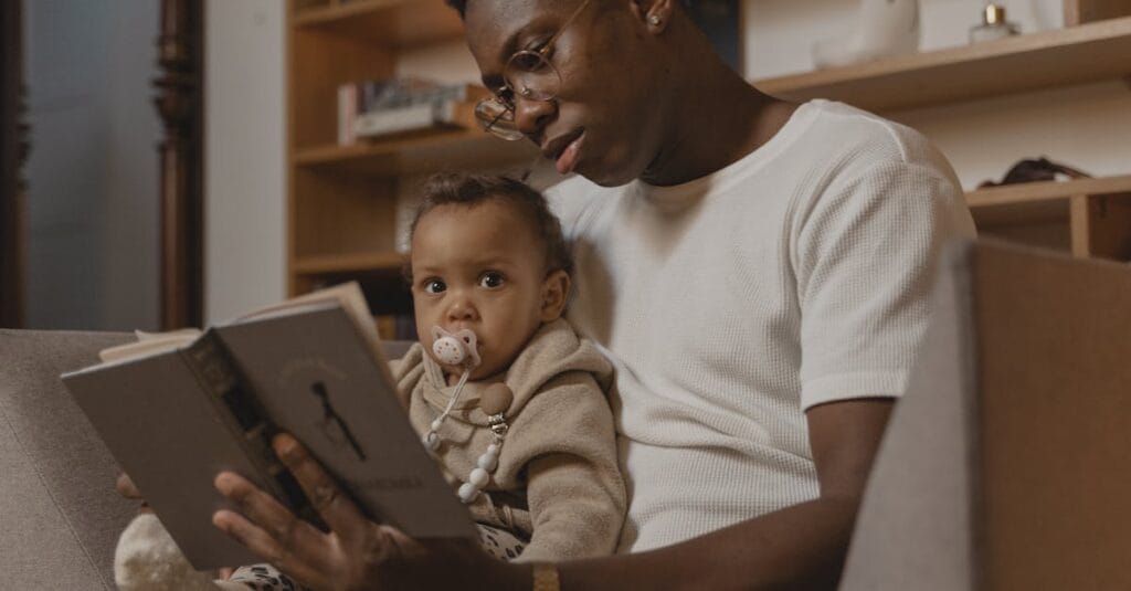 A father reads a book to his baby while sitting on a couch, creating a warm family moment.