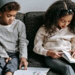 Grimacing stylish African American siblings reading books and sitting together on cozy couch in living room