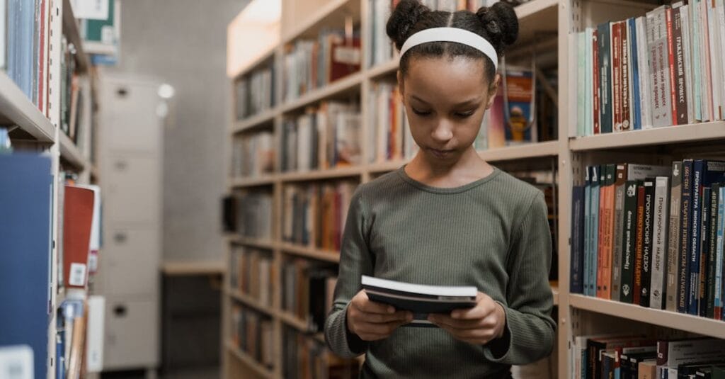 A young girl engrossed in reading a book while standing in a quiet library aisle.