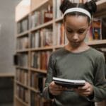 A young girl engrossed in reading a book while standing in a quiet library aisle.