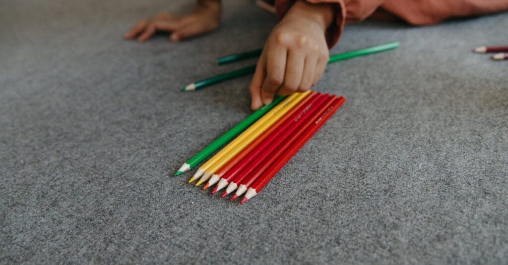 A hand organizes vibrant colored pencils on a carpeted floor.