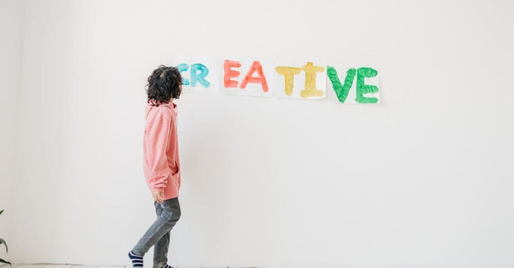 A child in casual attire creatively paints on a white wall indoors, spelling 'CREATIVE' with vivid colors.