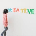 A child in casual attire creatively paints on a white wall indoors, spelling 'CREATIVE' with vivid colors.