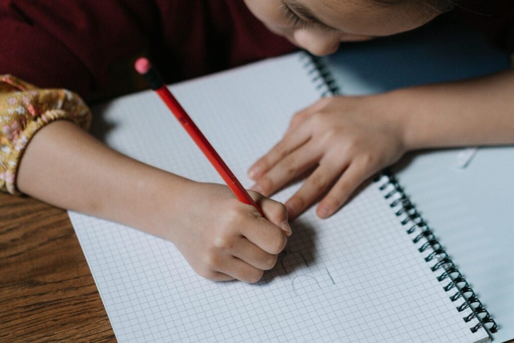 A child writes in a spiral notebook with a red pencil on a wooden desk.