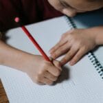 A child writes in a spiral notebook with a red pencil on a wooden desk.