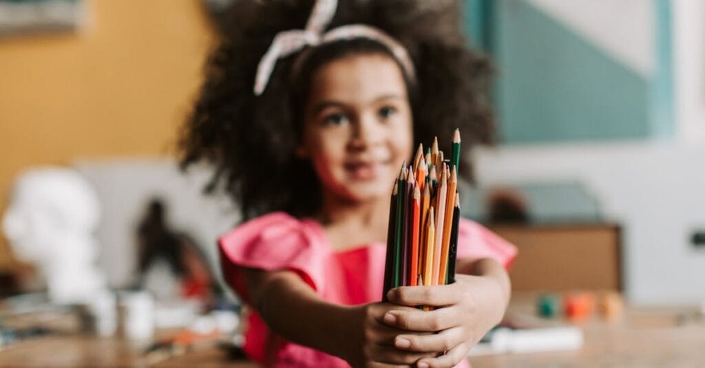 Smiling young girl in pink dress holding colored pencils, showcasing creativity in an art room.