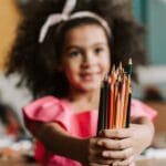 Smiling young girl in pink dress holding colored pencils, showcasing creativity in an art room.