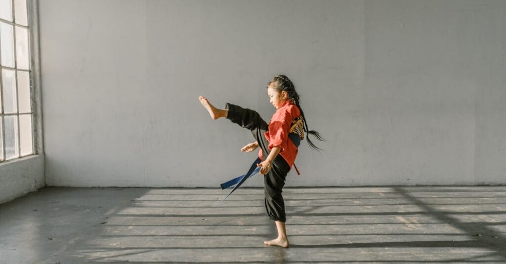 A young girl practicing a martial arts kick in a sunlit dojo indoor environment.