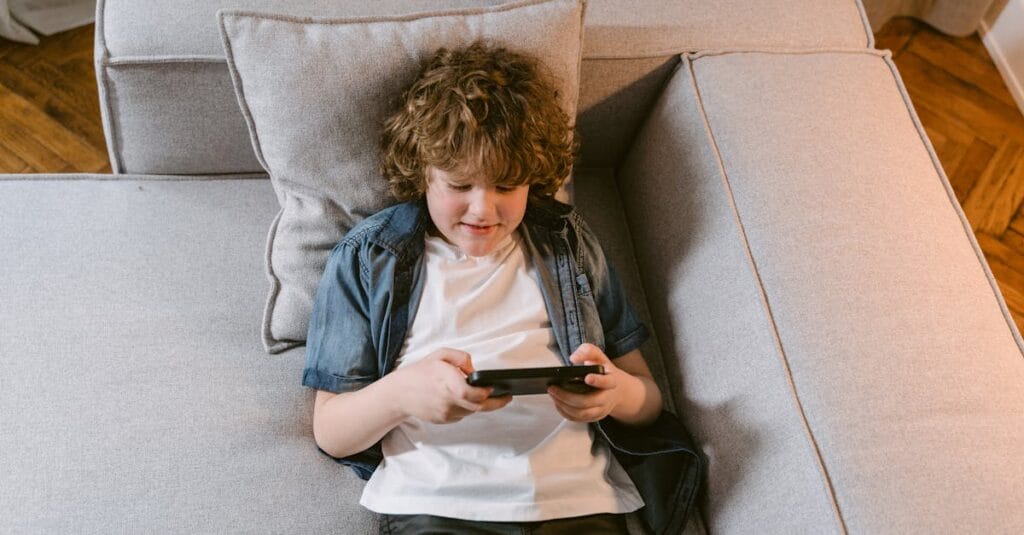Young boy lying on sofa playing with a mobile device, smiling and relaxed indoors.