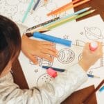 A child with pigtails coloring space-themed drawings with colorful pencils indoors.
