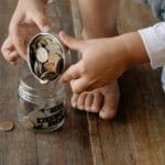 A child adds coins into a glass jar labeled for savings on a wooden floor.