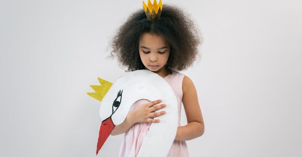 Cute African American child in pink dress and crown holding paper swan with crown while looking down and standing on white background in studio