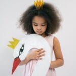 Cute African American child in pink dress and crown holding paper swan with crown while looking down and standing on white background in studio