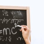 Close-up of hand writing physics equations on a chalkboard indoors.