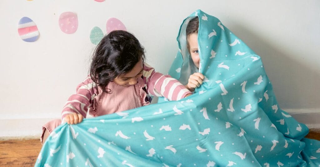 Two children playfully hiding under a colorful blanket indoors, surrounded by Easter decorations.