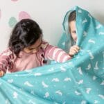 Two children playfully hiding under a colorful blanket indoors, surrounded by Easter decorations.
