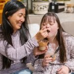 A mother joyfully feeds her daughter with Down syndrome on a sunny patio, sharing a snack together.