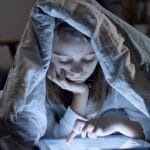A child using a tablet under a blanket in a dark room, creating a cozy bedtime atmosphere.