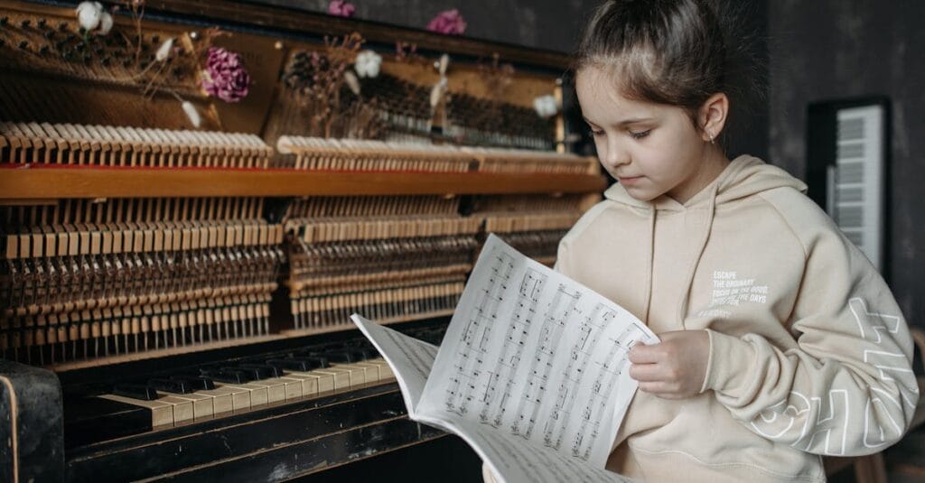 A young girl in a hoodie jacket sits by a piano, flipping through music sheets with focus.