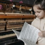 A young girl in a hoodie jacket sits by a piano, flipping through music sheets with focus.
