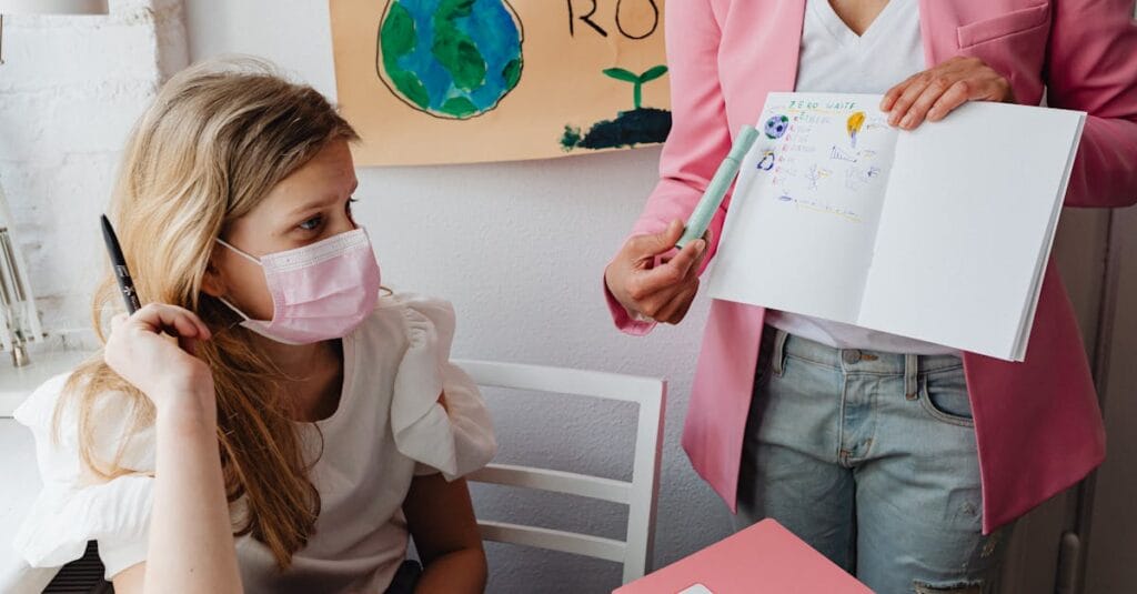 Young student wearing a facemask listens in class with a teacher showing a notebook.