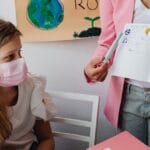 Young student wearing a facemask listens in class with a teacher showing a notebook.