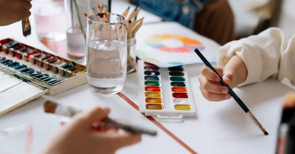 Children's hands holding paintbrushes while painting with watercolors during a creative art session.