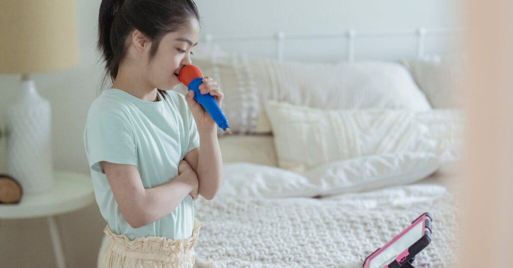 A young girl sings into a toy microphone while watching a tablet in her cozy bedroom.