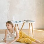 A young girl engaged in drawing on the wooden floor in a cozy, sunlit room.