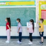 Teacher and diverse children wearing masks in line for classroom activity, embracing new normal.