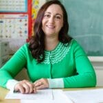 Engaging image of a smiling teacher seated at a desk in a classroom environment.
