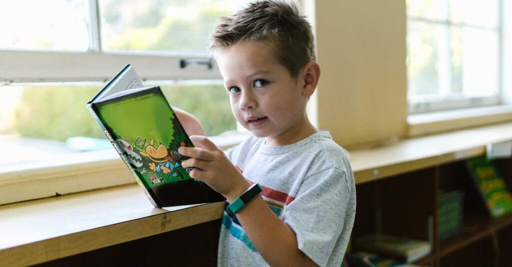 Young boy holding a colorful book while standing by a window, enjoying reading time.