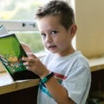 Young boy holding a colorful book while standing by a window, enjoying reading time.