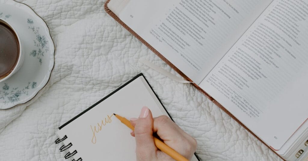 A hand writing in a notebook next to an open Bible and a cup of coffee on a quilted blanket.