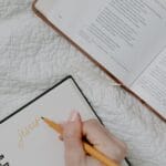 A hand writing in a notebook next to an open Bible and a cup of coffee on a quilted blanket.