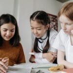 Three diverse schoolgirls working together over books in a classroom setting, fostering education and collaboration.