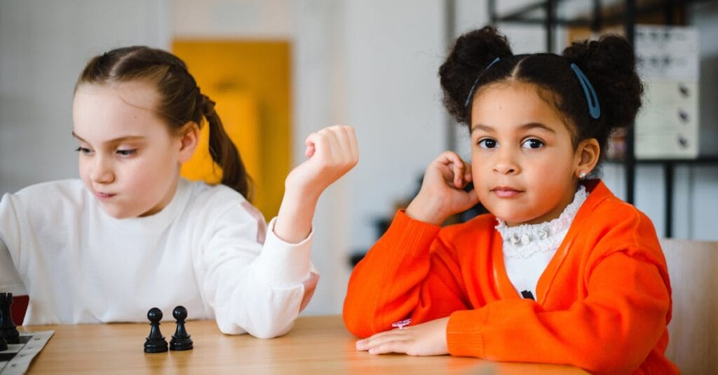 Two young girls engaged in a playful chess game indoors, showcasing focus and friendship.