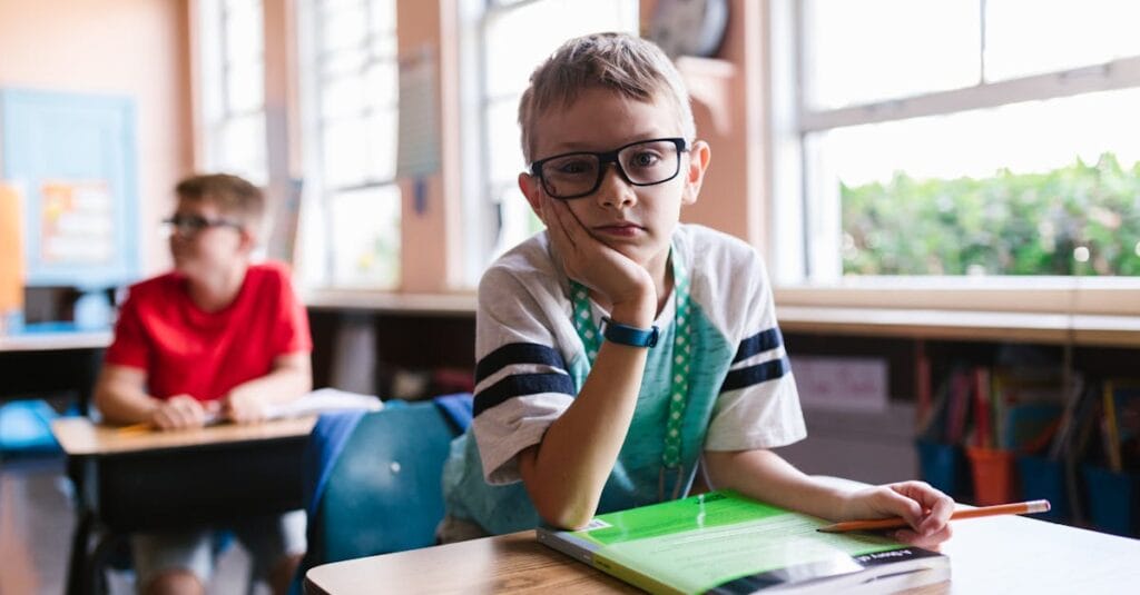 Young boy with glasses in a classroom, pen in hand, looking thoughtful.