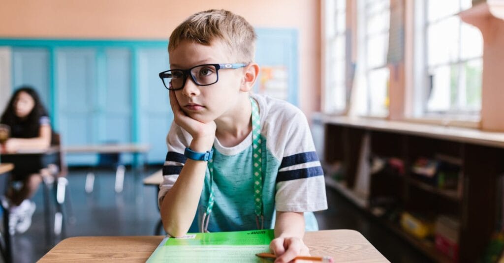 A young boy wearing eyeglasses sits at a desk in a classroom, focused on a book.