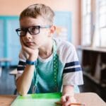 A young boy wearing eyeglasses sits at a desk in a classroom, focused on a book.