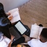 Three students sit on stairs, using laptops and notebooks, engaged in collaborative learning.