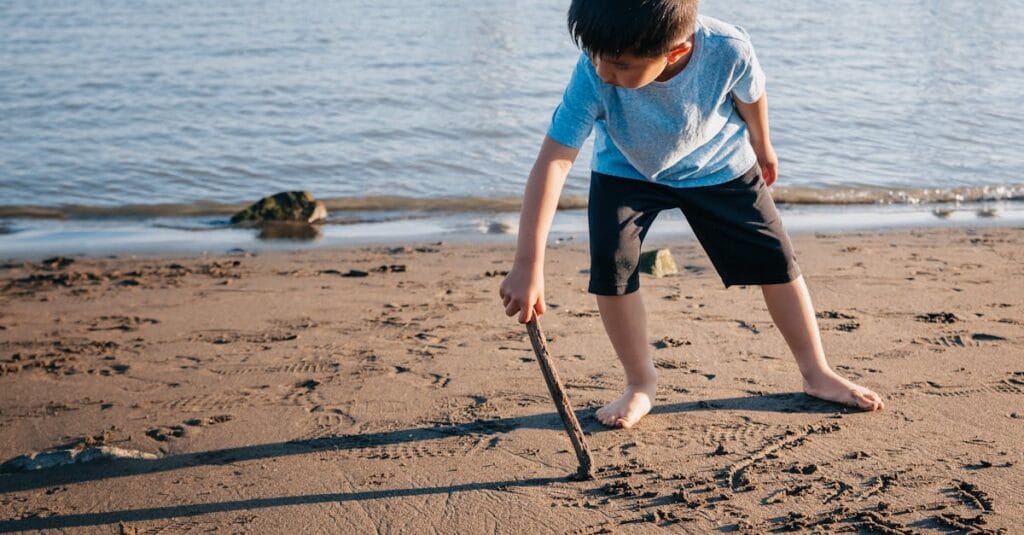 A young boy plays at the beach, drawing shapes in the sand with a stick.