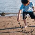 A young boy plays at the beach, drawing shapes in the sand with a stick.